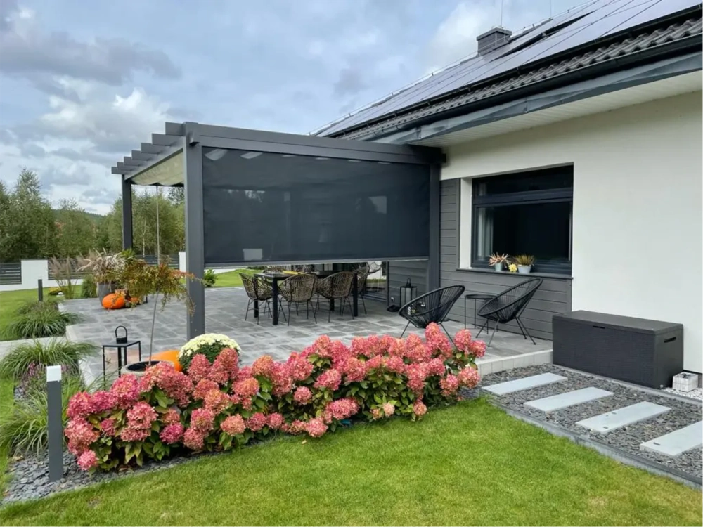 Zadaszenie na taras – aluminiowa pergola z boczną roletą, meble ogrodowe na kamiennym patio przy rabacie kwiatowej