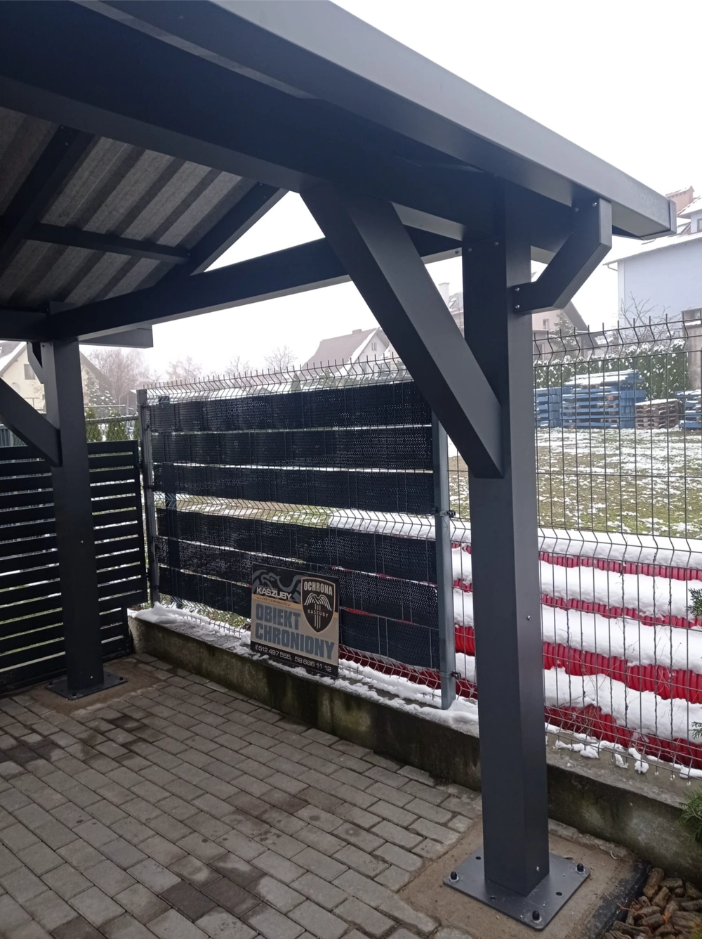 Aluminiowy carport z zadaszeniem z falistej blachy i słupami montowany przy ogrodzeniu