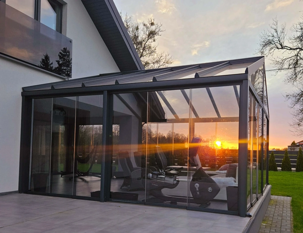 Ogrody zimowe z aluminiową konstrukcją i przeszklonym dachem na tarasie z rowerem stacjonarnym przy zachodzie słońca