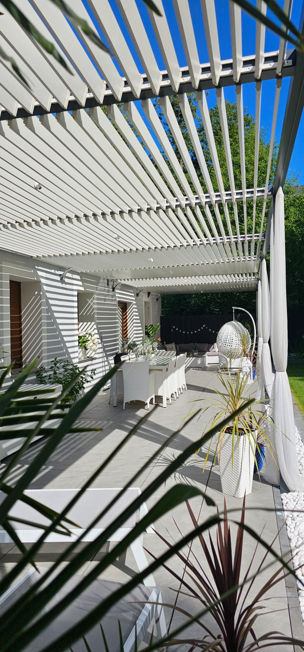Taras z białą pergolą z ruchomymi lamelami, dużym stołem jadalnym i fotelem bujanym – zadaszenia tarasowe